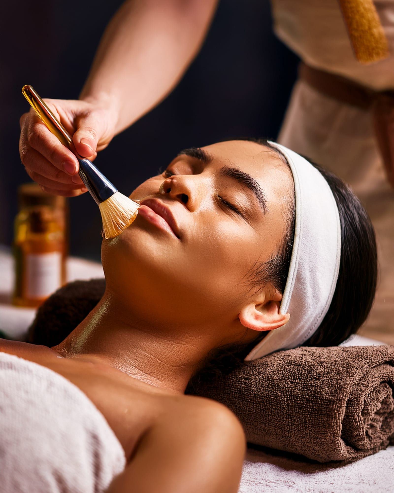 A fan brush laying a smooth cream mask along the cheek of a client lying back with her eyes closed.