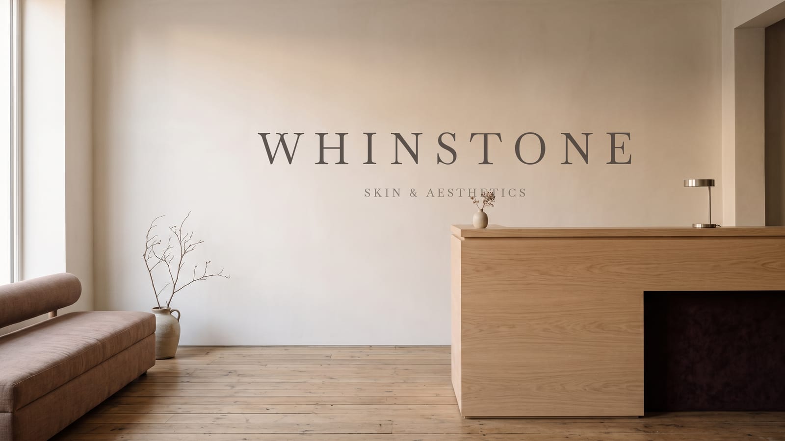 The WHINSTONE reception: a pale oak counter against a warm plaster wall, a low bench and a single bare branch in a stoneware vase.