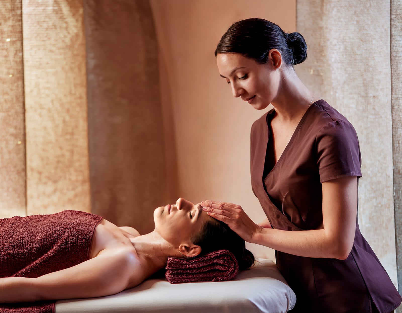 A therapist resting her fingertips at the temple of a client lying back with her eyes closed, in warm low light.